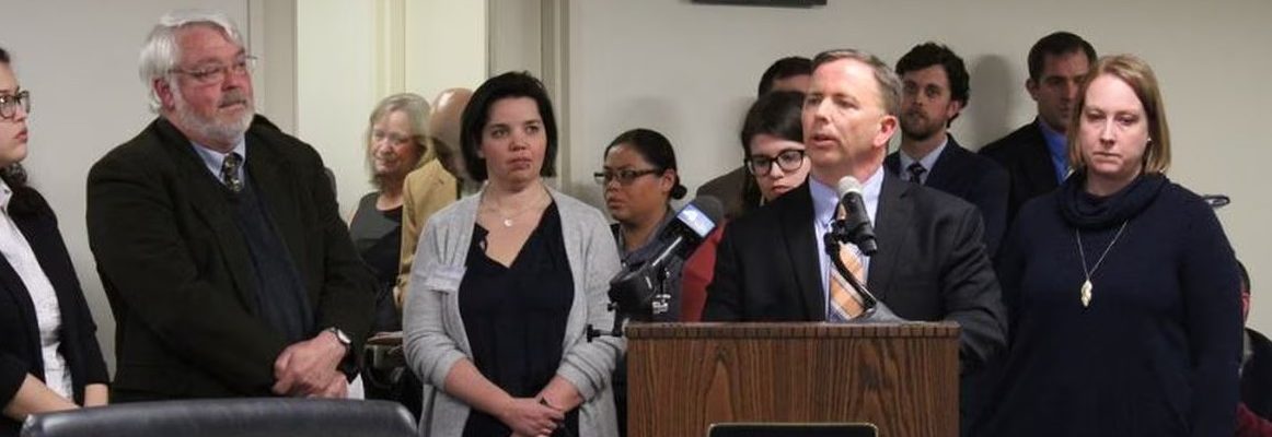 Frank Lomonte stands at a microphone with several other people listening nearby