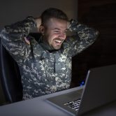 A man in a military uniform smiles at his laptop