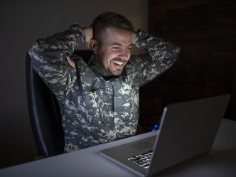 A man in a military uniform smiles at his laptop