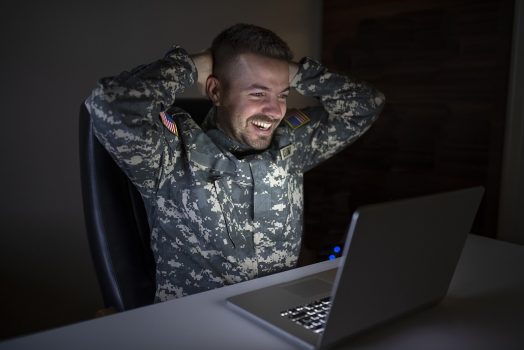 A man in a military uniform smiles at his laptop