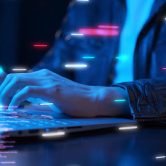 The hands of a cybersecurity professional work a keyboard to display lines of code, which are suspended in space around his laptop.