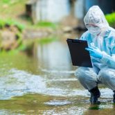 An environmental scientist crouches in a stream while holding a water sample and writing on a clipboard.
