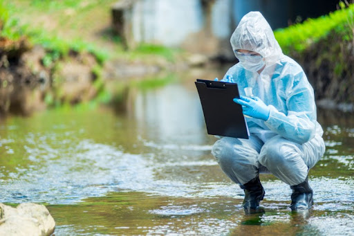 An environmental scientist crouches in a stream while holding a water sample and writing on a clipboard.