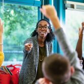 A teacher pointing to a group of students in class raising their hands.