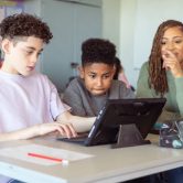 A teacher watches as students work on a tablet.