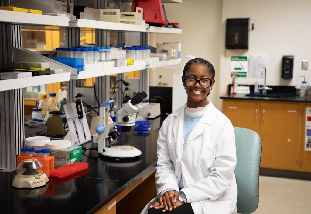 Student in white coat smiling next to lab equipment