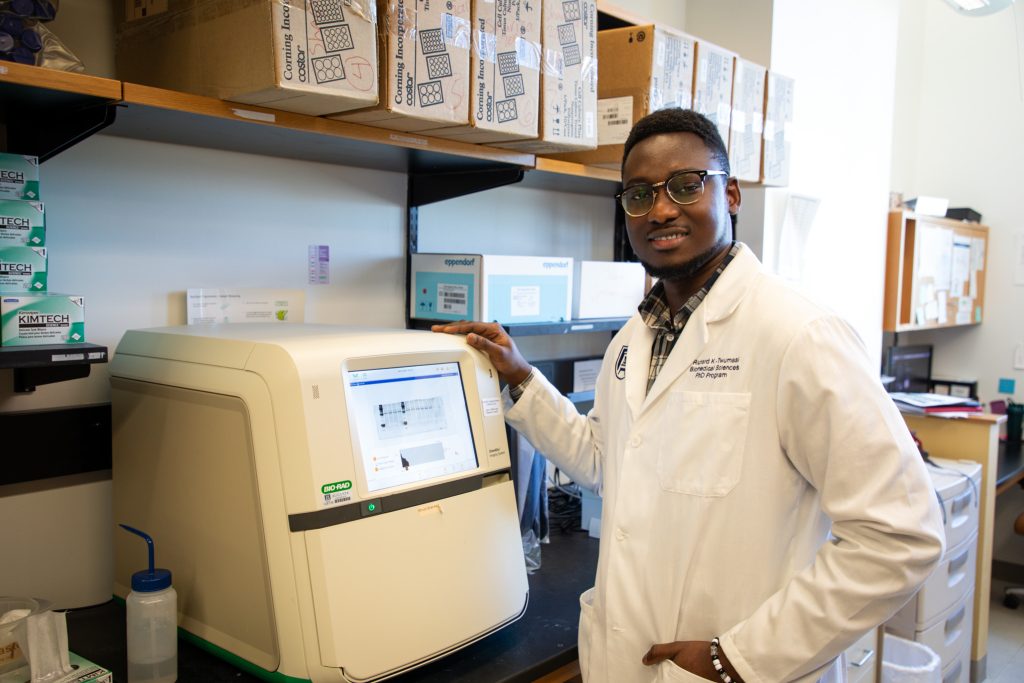 Student in white coat stands smiling next to lab equipment.