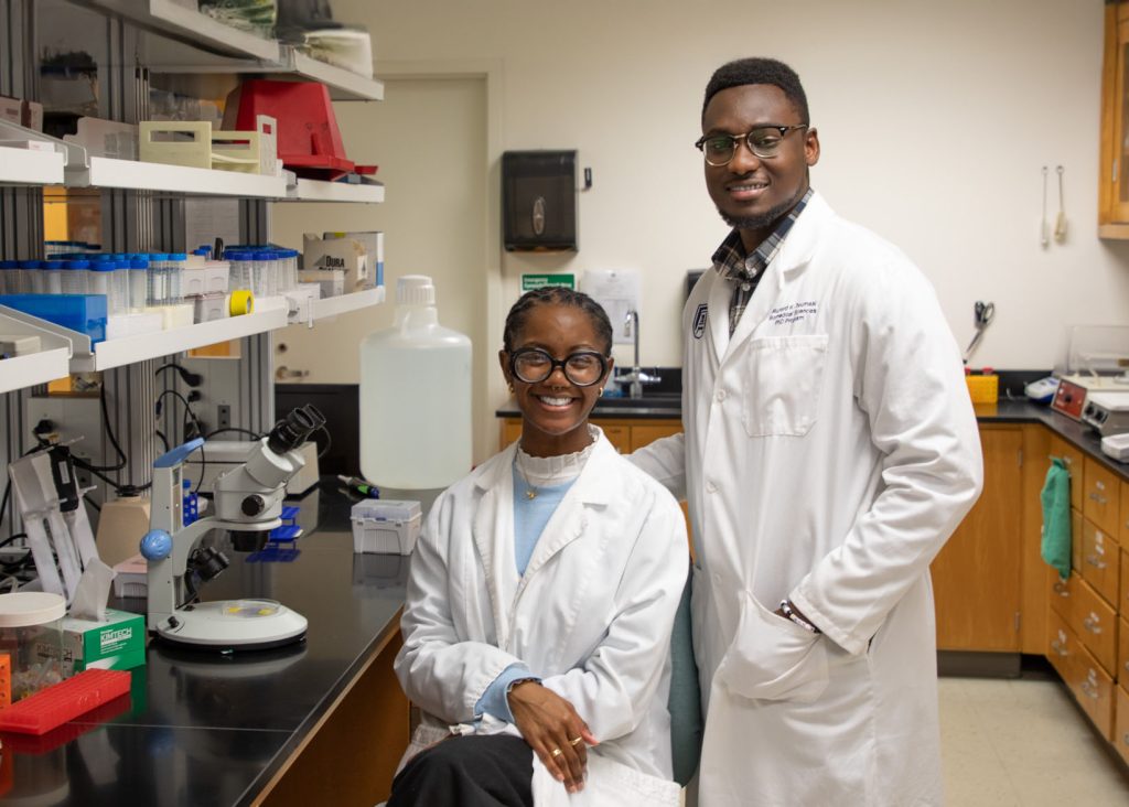 Two students wearing white coats posing in a research lab.
