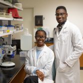 Two students wearing white coats posing in a research lab.