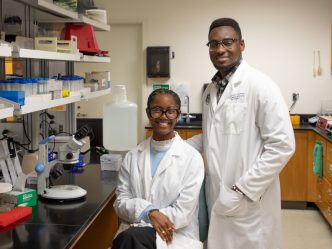 Two students wearing white coats posing in a research lab.