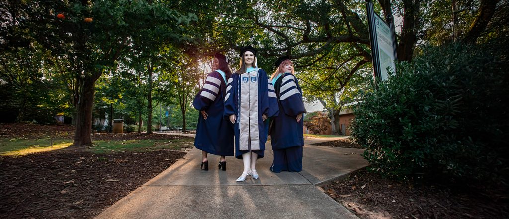 Three doctoral students stand in regalia