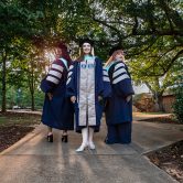 Three doctoral students stand in regalia