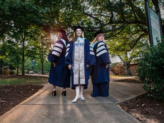 Three doctoral students stand in regalia