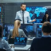 An intelligence director talks to a team of intelligence analysts working on their computers.