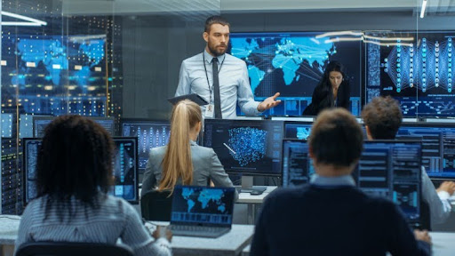 An intelligence director talks to a team of intelligence analysts working on their computers.