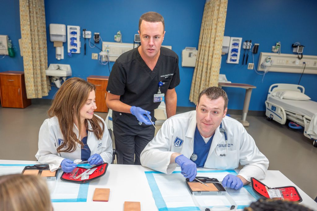 Students in white coats learn suturing from professor in gray scrubs