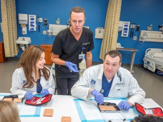 Students in white coats learn suturing from professor in gray scrubs