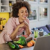 A health promotion specialist stands behind a counter with fruits and vegetables leading an online health education class.