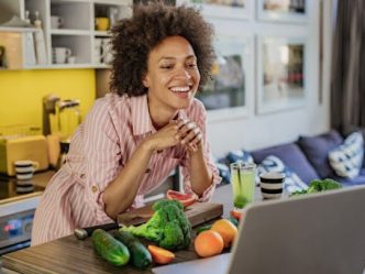 A health promotion specialist stands behind a counter with fruits and vegetables leading an online health education class.