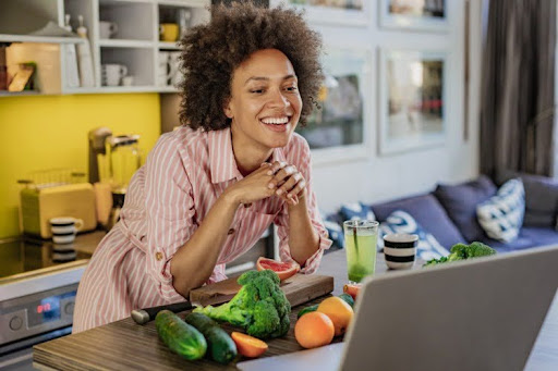 A health promotion specialist stands behind a counter with fruits and vegetables leading an online health education class.