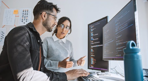 Two cybersecurity analysts working at a desk with several computers.