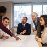 A city manager points to a map during a council meeting around a conference table.