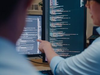 A cybersecurity manager points to an employee’s code on a monitor.