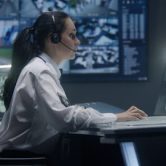 An immigration security officer wearing a headset watches surveillance footage on a computer.