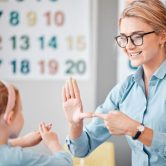 A smiling teacher uses sign language with a child in a classroom.