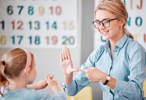 A smiling teacher uses sign language with a child in a classroom.