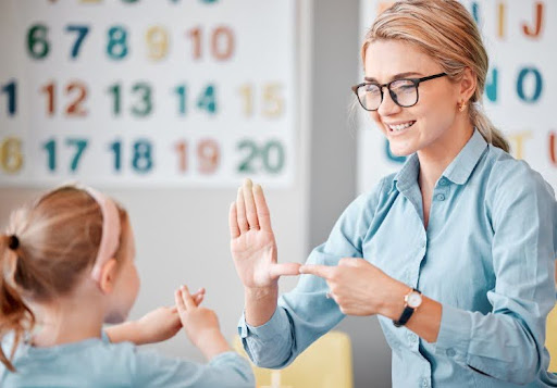 A smiling teacher uses sign language with a child in a classroom.