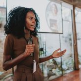 A training and development specialist holds a microphone and speaks in front of a projector screen in an office.