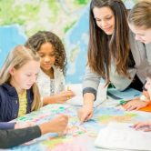 A social studies teacher and several students gather around a table and examine a world map.