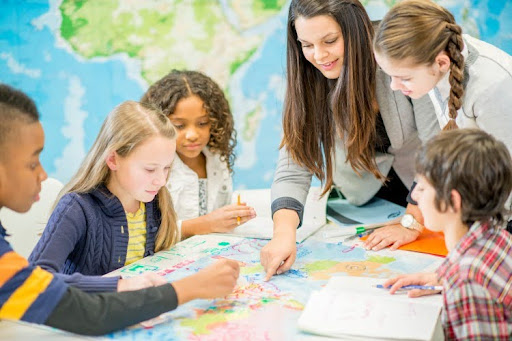 A social studies teacher and several students gather around a table and examine a world map.