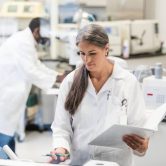 A clinical research coordinator holding a binder enters information into a computer in a lab.