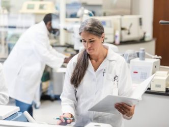 A clinical research coordinator holding a binder enters information into a computer in a lab.