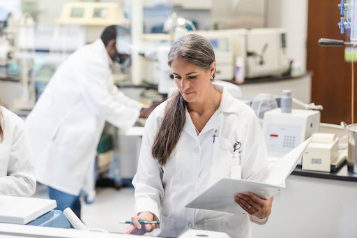 A clinical research coordinator holding a binder enters information into a computer in a lab.