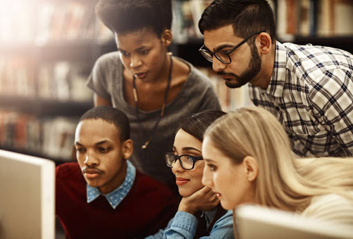 A group of five students gather around a computer in a library.