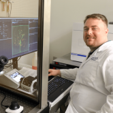 Corey Andrews smiles at the camera, wearing a lab coat and sitting in front of a computer.