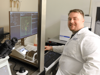 Corey Andrews smiles at the camera, wearing a lab coat and sitting in front of a computer.