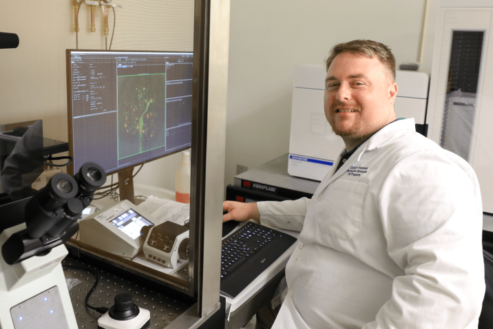 Corey Andrews smiles at the camera, wearing a lab coat and sitting in front of a computer.