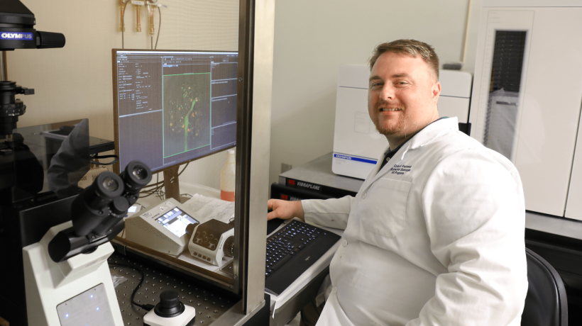 Corey Andrews smiles at the camera, wearing a lab coat and sitting in front of a computer.