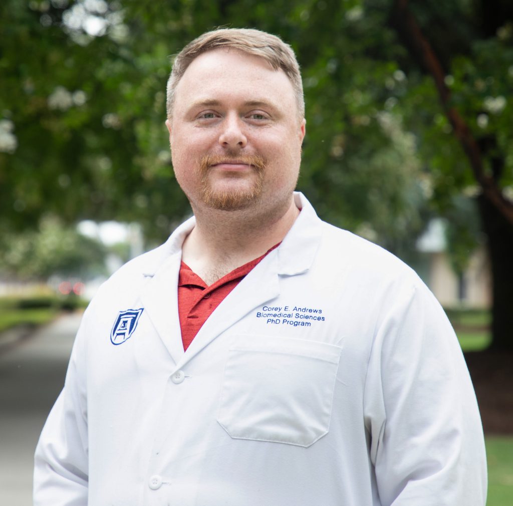 Headshot of Corey Andrews smiling at the camera, wearing his white lab coat, standing on the sidewalk in front of greenery.