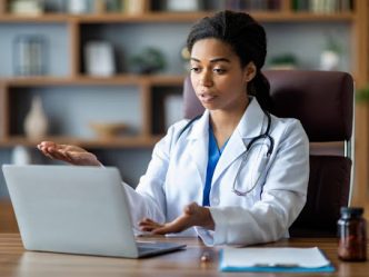 A telehealth care provider wearing a white coat talks with a patient using a laptop.