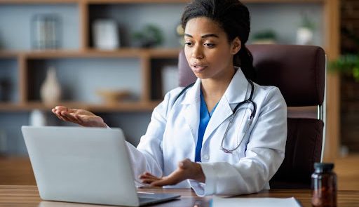 A telehealth care provider wearing a white coat talks with a patient using a laptop.