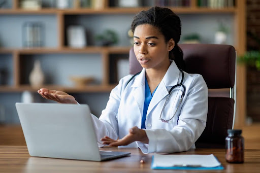 A telehealth care provider wearing a white coat talks with a patient using a laptop.